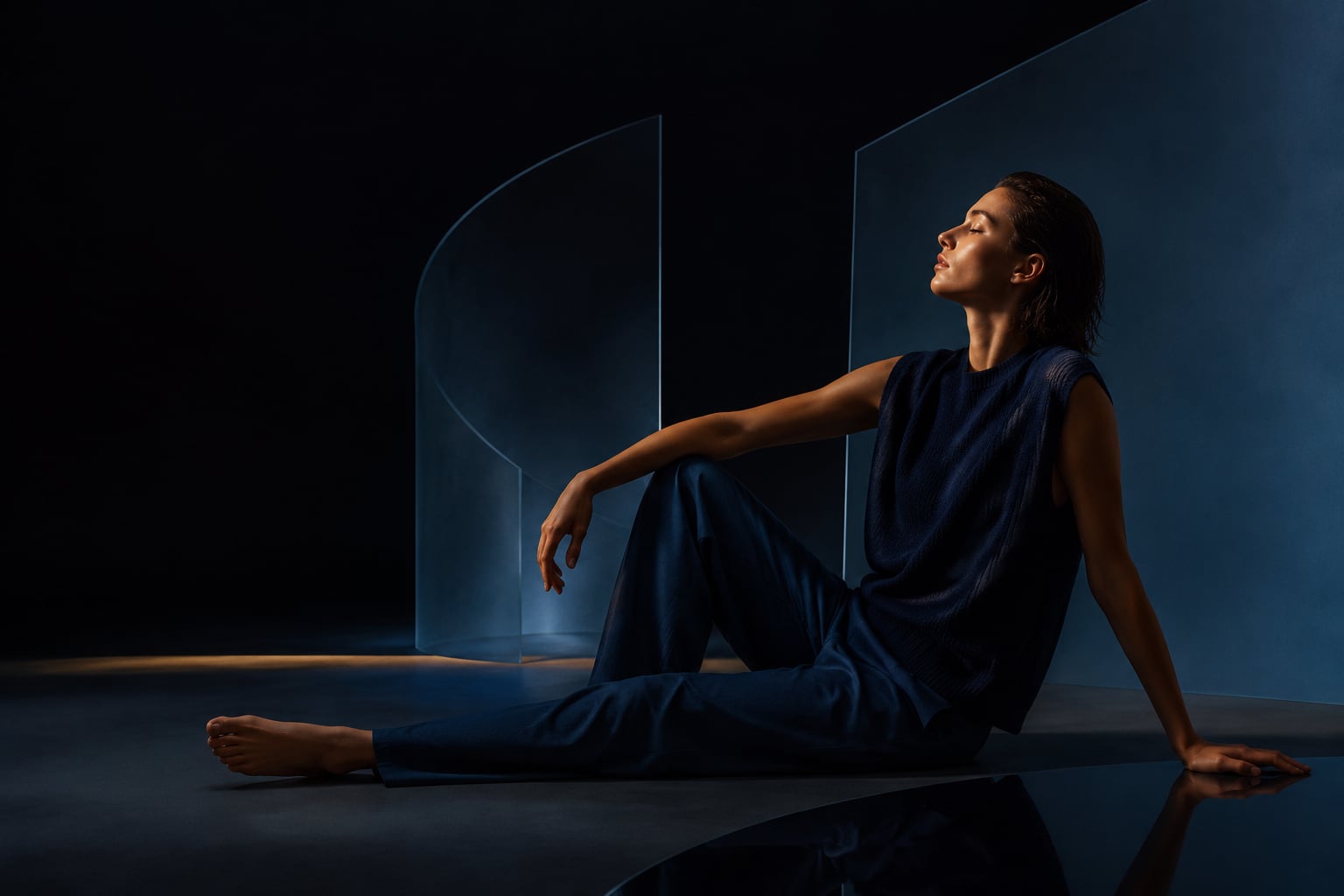 A woman in deep navy clothing seated in a sculptural blue room, held by a soft warm edge of light