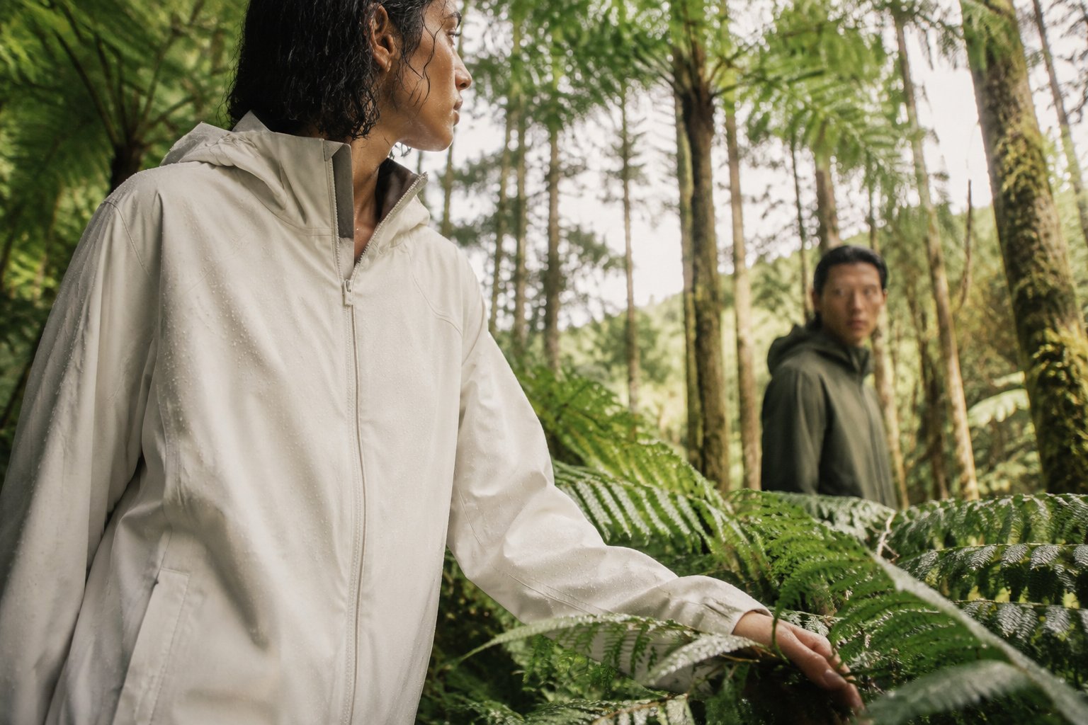 A person in a rain jacket reaches through wet ferns while a second person stands behind in the forest